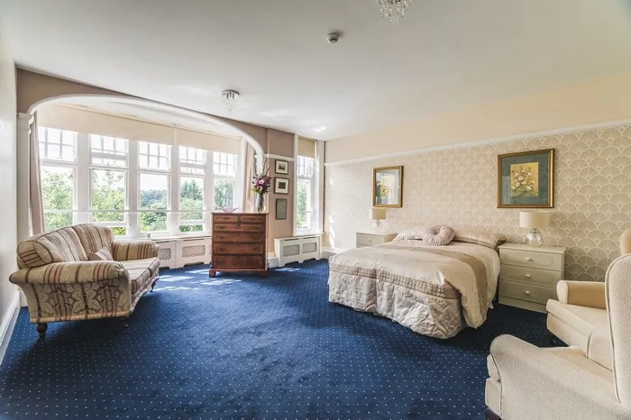 A double bedroom with a blue carpet, a sofa and armchairs beside a large window